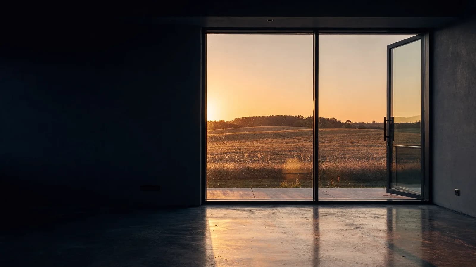 Low sun streaming through a floor-to-ceiling glass wall into a dark room with a polished concrete floor